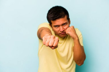 Latin kökenli genç bir adam mavi arka planda izole edilmiş. Yumruk atıyor, sinirleniyor, kavga ediyor..