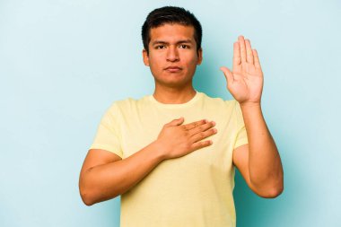 Genç İspanyol bir adam mavi arka planda izole edilmiş yemin ediyor, elini göğsüne koyuyor..