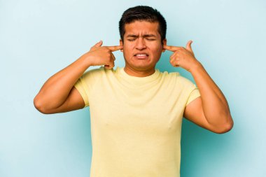 Genç İspanyol bir adam mavi arka planda izole edilmiş parmaklarıyla kulaklarını kapatıyor, gürültülü bir ortam yüzünden stresli ve çaresiz..