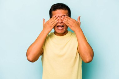 Mavi arka planda izole edilmiş genç İspanyol adam elleriyle gözlerini kapatıyor, geniş bir gülümsemeyle bir sürpriz bekliyor..