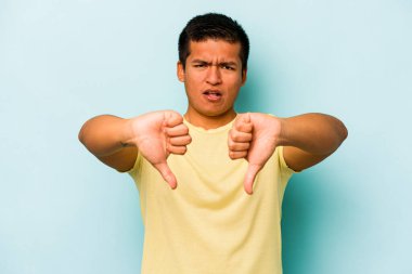 Mavi arka planda izole edilmiş genç İspanyol adam baş parmağını aşağı gösteriyor ve hoşlanmadığını ifade ediyor..