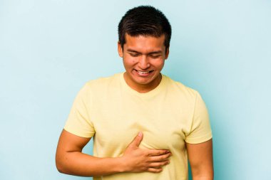 Mavi arka planda izole edilmiş genç İspanyol adam karnına dokunuyor, nazikçe gülümsüyor, yemek yiyor ve tatmin oluyor..