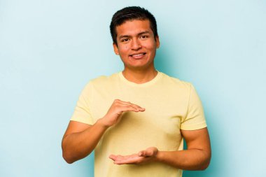 Mavi arka planda izole edilmiş genç İspanyol adam iki eliyle bir şey tutuyor, ürün sunumu..