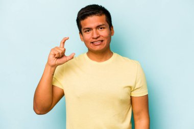 Mavi arka planda izole edilmiş genç İspanyol adam işaret parmaklarıyla küçük bir şey tutuyor, gülümsüyor ve kendinden emin..