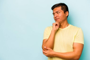 Genç İspanyol adam mavi arka planda izole edilmiş şüpheli ve şüpheci bir ifadeyle yana bakıyor..