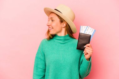 Pembe arka planda izole edilmiş pasaport taşıyan beyaz bir kadın gülümsüyor ve keyifli görünüyor..