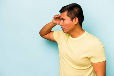 Genç İspanyol adam mavi arka planda izole edilmiş, uzaklara bakıyor ve elini alnına koyuyor..