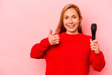 Pembe arka planda izole edilmiş genç beyaz kadın gülümsüyor ve başparmağını kaldırıyor.