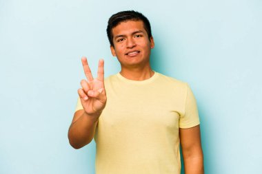 Mavi arka planda izole edilmiş genç İspanyol adam zafer işareti gösteriyor ve gülümsüyor..