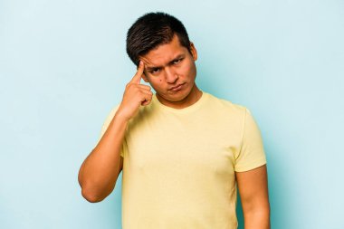 Genç İspanyol bir adam mavi arka planda izole edilmiş, parmağıyla tapınağı işaret ediyor, düşünüyor ve bir göreve odaklanıyor..