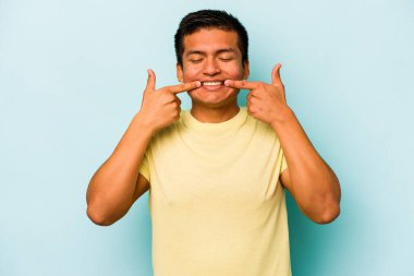 Mavi arka planda izole edilmiş genç İspanyol adam gülümsüyor, parmaklarını ağıza doğrultuyor..