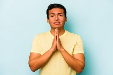 Mavi arka planda izole edilmiş genç bir İspanyol. El ele tutuşup, ağzının kenarında dua ediyor. Kendine güveniyor..