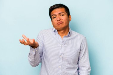 Latin kökenli genç bir adam, mavi arka planda izole edilmiş şüphe içinde ve sorgulama sırasında omuz silkiyor..