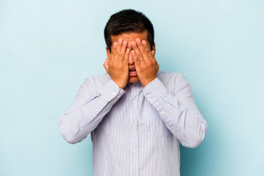 Genç İspanyol adam mavi arka planda izole edilmiş. Yüzünün yarısını avuç içiyle kaplayarak eğleniyor..