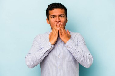 Mavi arka planda izole edilmiş genç İspanyol adam elleriyle ağzını kapatırken şok oldu..