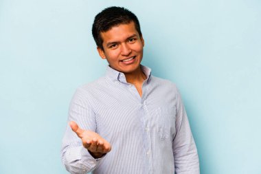 Mavi arka planda izole edilmiş genç İspanyol adam selamlama jesti olarak kameraya uzanıyor..