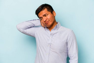 Genç İspanyol adam mavi arka planda izole edilmiş. Çok yorgun ve uykulu. Elini başının üstünde tutuyor..