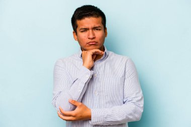Genç İspanyol bir adam mavi arka planda izole edilmiş şüpheli, belirsiz, seni muayene ediyor..