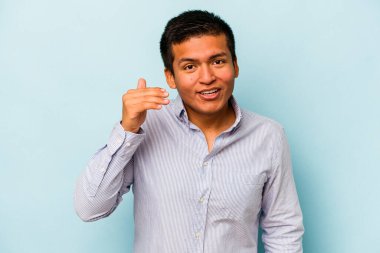 Genç İspanyol bir adam mavi arka planda izole edilmiş bir şekilde bir şeye gülüyor, elleriyle ağzını kapatıyor..