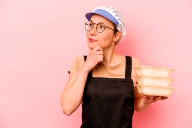 Pembe arka planda izole edilmiş genç aşçı kadın şüpheli ve şüpheci bir ifadeyle yana bakıyor..