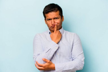 Latin asıllı genç adam mavi arka planda izole edilmiş. Mutsuz bir şekilde kameraya bakıyor. İğneleyici bir ifadeyle..