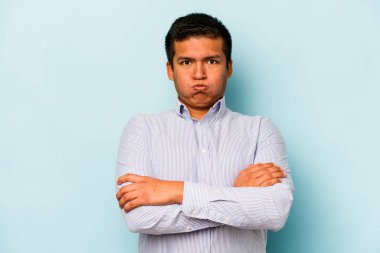 Mavi arka planda izole edilmiş genç İspanyol adam yanakları patlatıyor, yorgun bir ifadesi var. Yüz ifadesi kavramı.