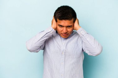 Genç İspanyol bir adam mavi arka planda izole edilmiş kulaklarını elleriyle kapatıyor çok fazla ses duymamaya çalışıyor..