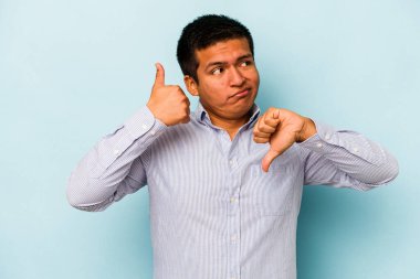 Latin kökenli genç adam mavi arka planda izole edilmiş, baş parmağını kaldırıyor, zor bir seçim konsepti.