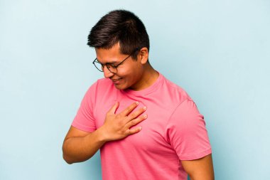 Mavi arka planda izole olmuş genç İspanyol adam gülüyor, ellerini yüreğine koyuyor, mutluluk kavramını koruyor..