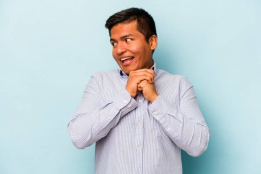 Mavi arka planda izole edilmiş genç İspanyol adam ellerini çenesinin altında tutuyor ve mutlu bir şekilde yan tarafa bakıyor..