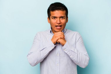 Latin kökenli genç adam mavi arka planda izole edilmiş şans için dua ediyor, şaşırmış ve ağzı açık bir şekilde önüne bakıyor..