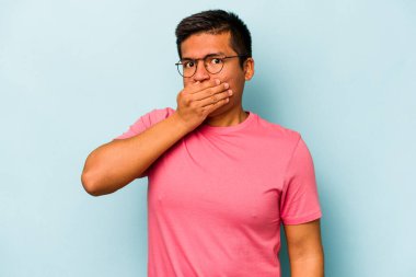 Genç İspanyol bir adam mavi arka planda izole edilmiş, ağzı kapalı ve elleri endişeli görünüyor..