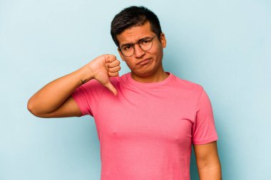 Mavi arka planda izole edilmiş genç İspanyol adam baş parmağını aşağı gösteriyor, hayal kırıklığı konsepti.