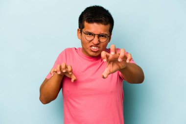 Mavi arka planda izole edilmiş genç İspanyol bir adam pençelerini bir kediyi taklit ederken gösteriyor, agresif bir hareket..