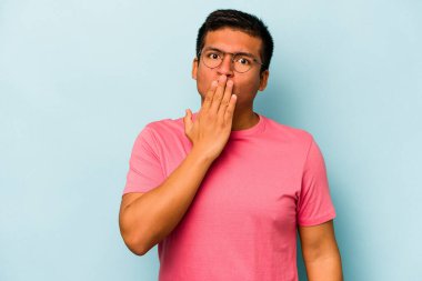 Mavi arka planda izole edilmiş genç İspanyol adam şok olmuş, ağzı ellerle kaplanmış, yeni bir şey keşfetmek için sabırsızlanıyor..