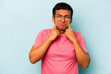 Genç İspanyol bir adam mavi arka planda izole edilmiş, ağlıyor, bir şeyden memnun değil, acı ve kafa karışıklığı konsepti..