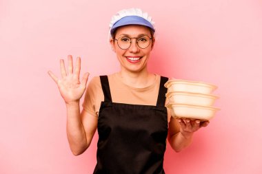 Genç aşçı gönüllü kadın pembe arka planda izole edilmiş gülümsüyor beş numarayı parmaklarıyla gösteriyor..