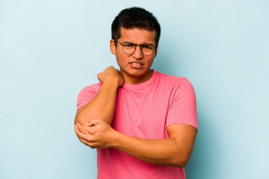 Genç İspanyol adam mavi arka planda izole edilmiş dirsek masajı yapıyor, kötü bir hareketin ardından acı çekiyor..