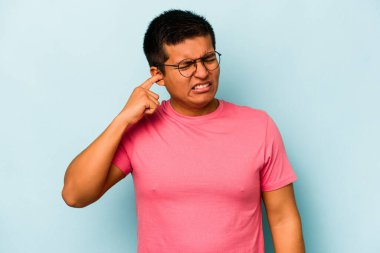 Genç İspanyol bir adam mavi arka planda izole edilmiş parmaklarıyla kulaklarını kapatıyor, gürültülü bir ortam yüzünden stresli ve çaresiz..