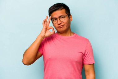 Mavi arka planda izole edilmiş genç İspanyol bir adam bir göz kırptı ve elleriyle iyi bir jest yaptı..