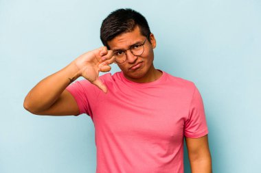 Mavi arka planda izole edilmiş genç İspanyol adam baş parmağını aşağı gösteriyor ve hoşlanmadığını ifade ediyor..