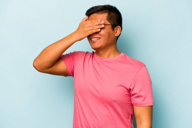 Mavi arka planda izole edilmiş genç İspanyol adam elleriyle gözlerini kapatıyor, geniş bir gülümsemeyle bir sürpriz bekliyor..