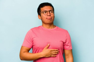 Mavi arka planda izole edilmiş genç İspanyol adam karnına dokunuyor, nazikçe gülümsüyor, yemek yiyor ve tatmin oluyor..