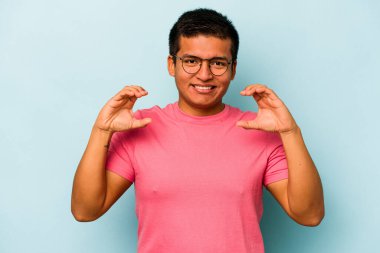 Genç İspanyol bir adam mavi arka planda izole edilmiş. Elinde avuç içi olan bir şeyle kameraya sunuyor..