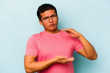 Mavi arka planda izole edilmiş genç İspanyol adam iki eliyle bir şey tutuyor, ürün sunumu..