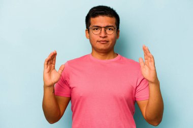 Mavi arka planda izole edilmiş genç İspanyol adam işaret parmaklarıyla küçük bir şey tutuyor, gülümsüyor ve kendinden emin..