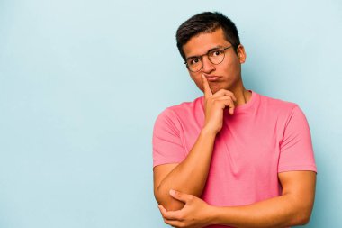Genç İspanyol adam mavi arka planda izole edilmiş şüpheli ve şüpheci bir ifadeyle yana bakıyor..