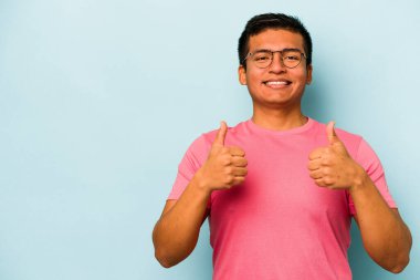 Genç İspanyol adam mavi arka planda izole edilmiş gülümsüyor ve başparmağını kaldırıyor.