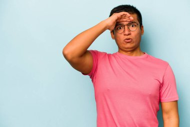 Genç İspanyol adam mavi arka planda izole edilmiş, uzaklara bakıyor ve elini alnına koyuyor..