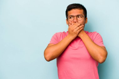 Genç İspanyol adam mavi arka planda izole edilmiş. Düşünceli bir şekilde bir fotokopi odasına bakıyor. Ağzını eliyle kapatıyor..